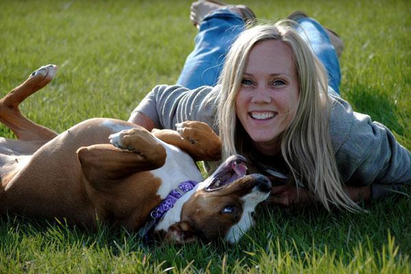 A woman lays on the grass with a brown and white dog laying on its back beside her.