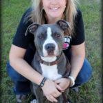 A woman sits with a grey and white dog. They are both looking at the camera.
