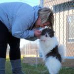 A woman bends over to hold the paws of a small fluffy black and white dog standing on two legs.