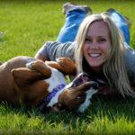 A woman lays on the grass with a brown and white dog mid-roll beside her.