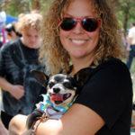 A woman with curly hair and red sunglasses smiles at the camera while holding a small dog in her arms.