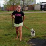 A woman wearing a black NDSU t-shirt smiles wide while accompanied by a small white dog on a leash.