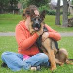 A woman sits beside a German Shepherd with her arms around him in a hug.