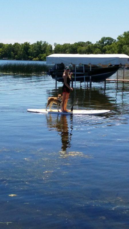 A woman and brown dog stand on a paddle board in the middle of a lake
