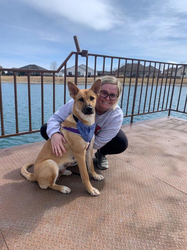 A woman kneels down beside a brown dog. They are on a dock with a lake in the background.