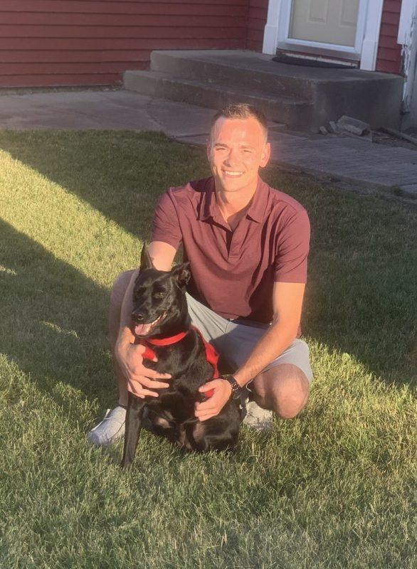 A man kneels with a small black dog with its tongue out.