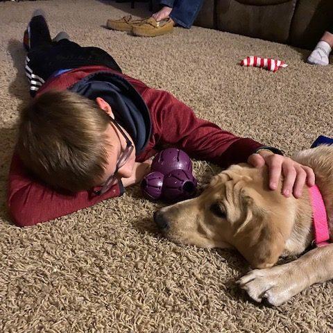 A tan dog lays with its head on the floor, next to a young boy. They are staring closely at each other. The young boy is petting the dog's head.