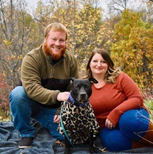 A man and woman pose in front of autumn foliage. A black dog wearing a leopard print sweater sits between them.