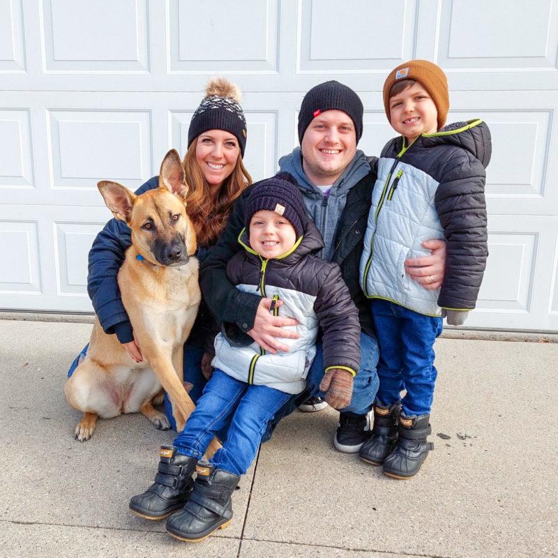 A family of four (a man, a woman, and two children) pose outside with a brown dog with very perky ears.