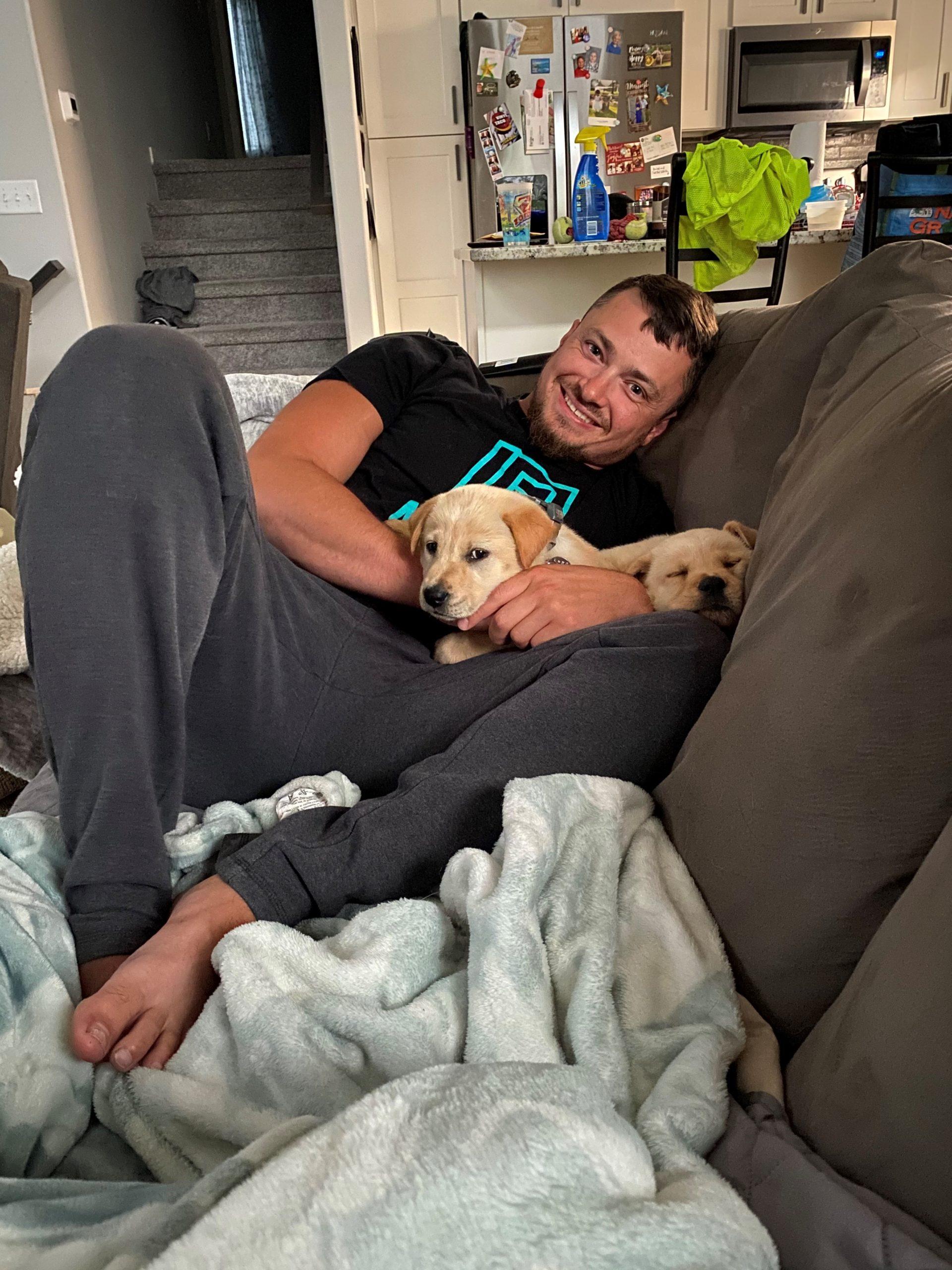 A man lays on a couch, smiling, while two golden retriever puppies sleep on him.