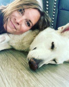 A woman smiles while resting her head on a sleeping white dog.