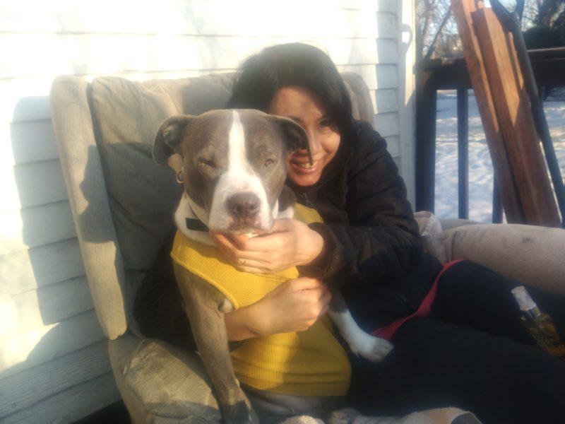 A young girl hugs a grey and white dog. They are on a chair outside, and the dog is wearing a yellow sweater.