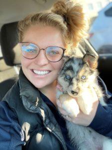 A woman with blonde hair in a big bun poses with an Australian Shepherd puppy.