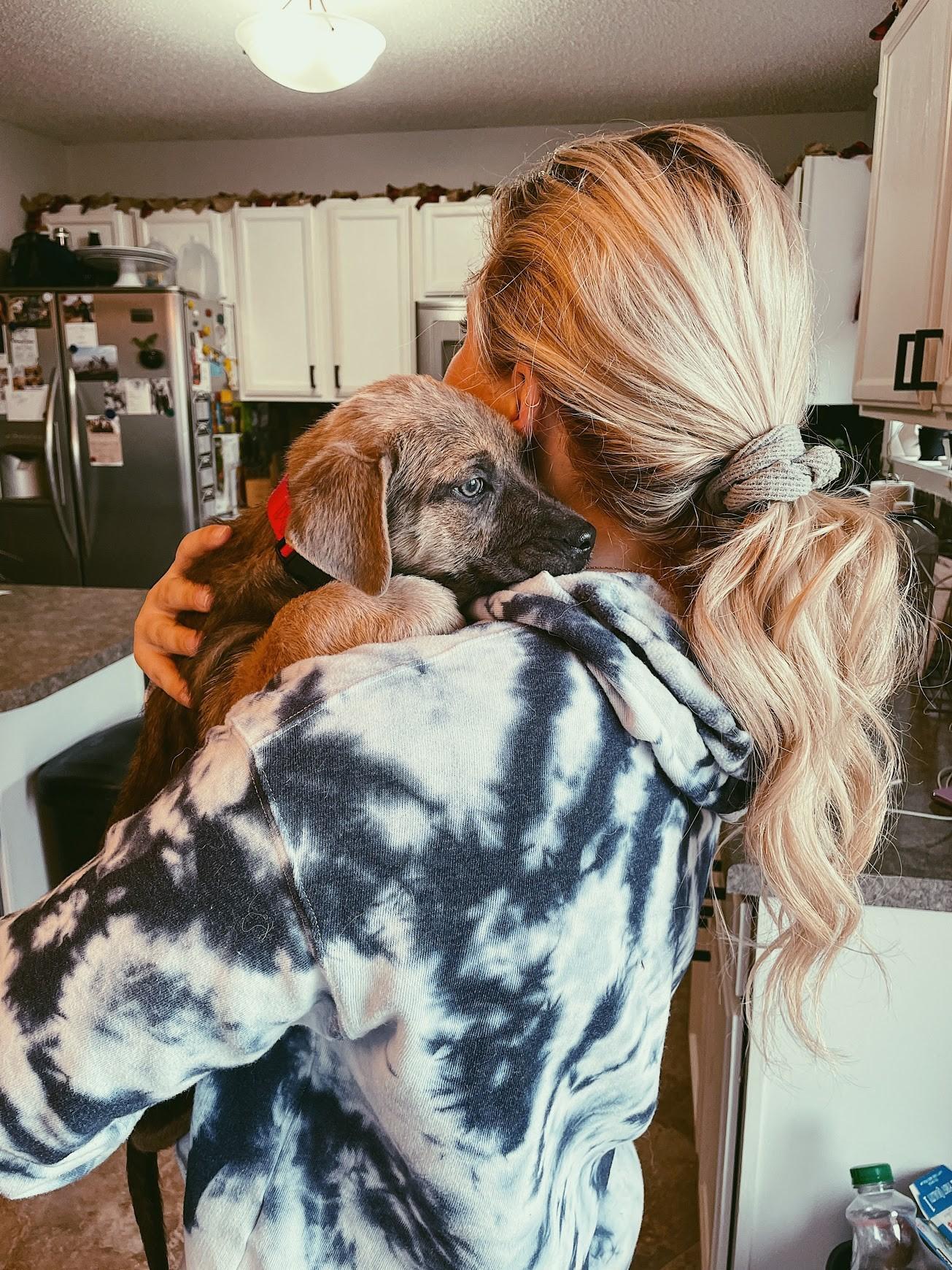 A woman with long blonde hair holds a brown puppy over her shoulder. The picture is taken from the back of the woman.
