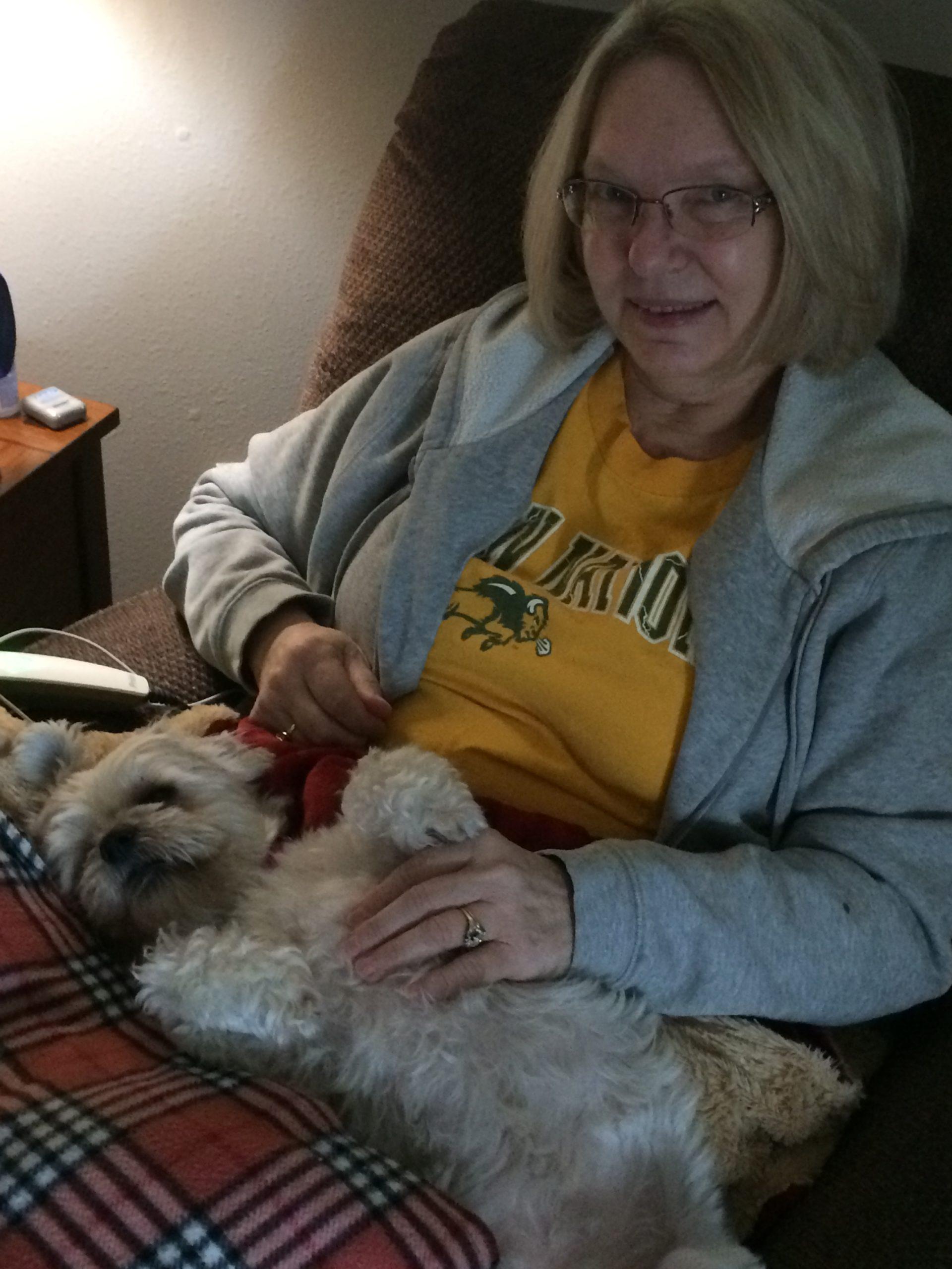 Foster Maran sits with a checkered blanket and a small white dog in her lap.