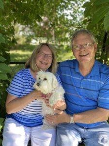 Foster Maran sits with her husband under some trees. Maran holds a small white dog.