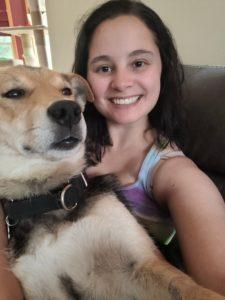 A woman takes a selfie while wearing a big smile. There is a brown dog in her lap.