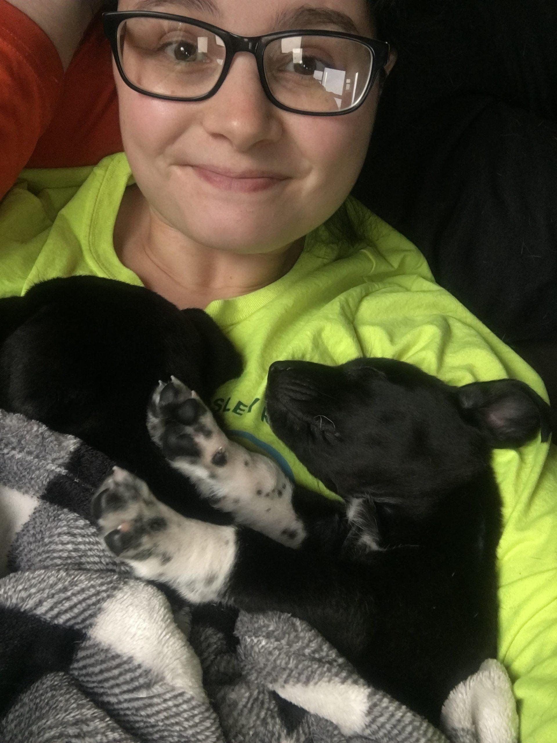 A woman in a neon green hoodie takes a selfie with two black puppies asleep on her.