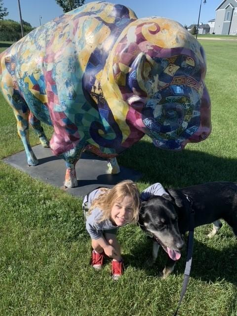 A little girl posts with a black dog in front of a North Dakota Bison sculpture.