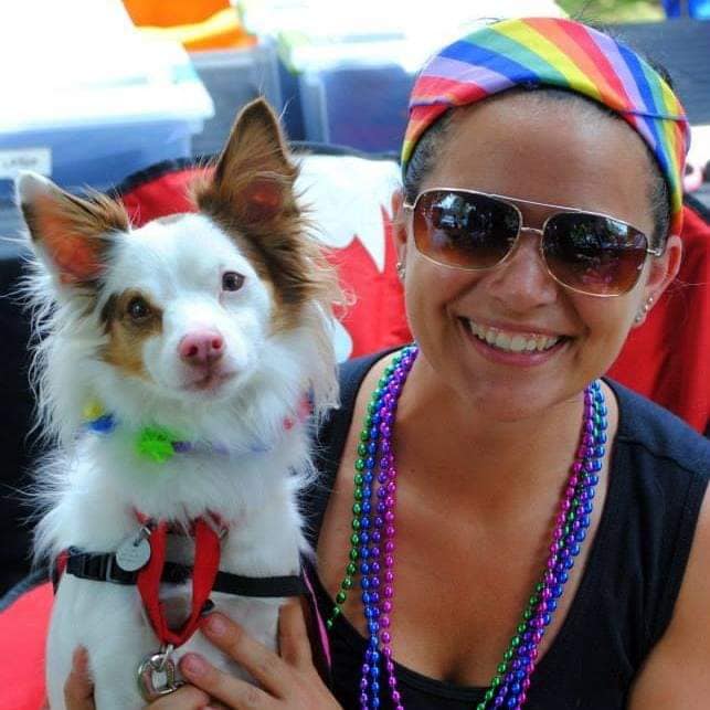 A woman wearing rainbow colors smiles while holding a small white dog with brown perky ears.