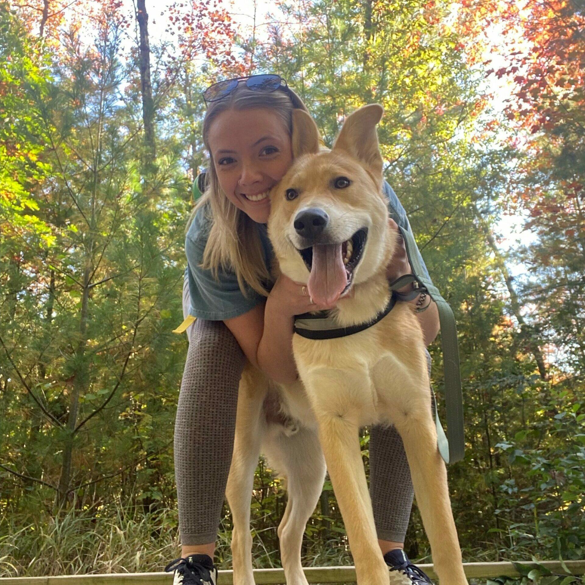 A woman poses outside with a big smiling yellow dog