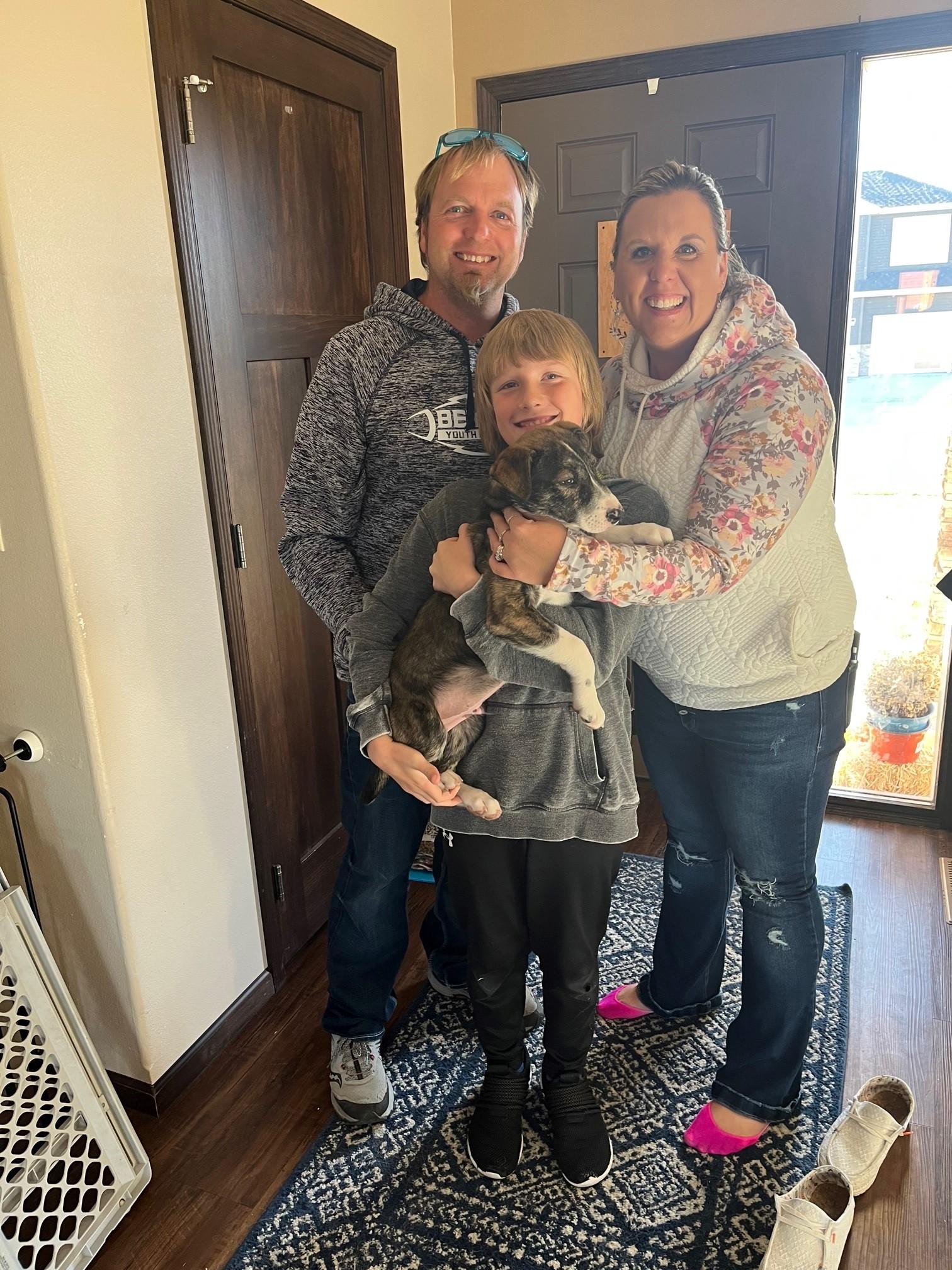 A family consisting of a father, mother, and child pose together in a home's entryway. They are smiling widely, and the child holds a small dog in his arms.